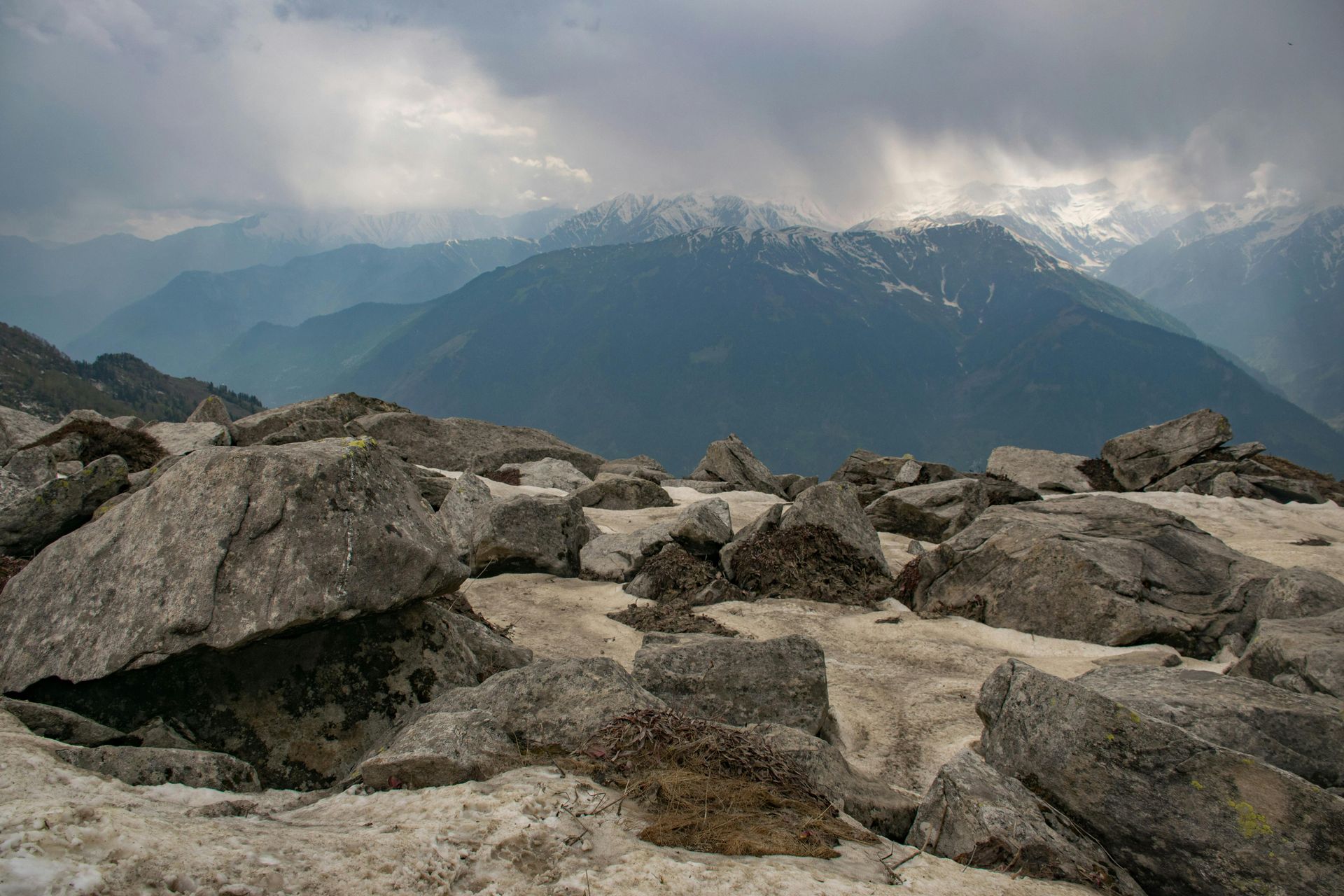 Rocky mountaintop with snow patches, overlooking a vast mountain range under a cloudy sky.