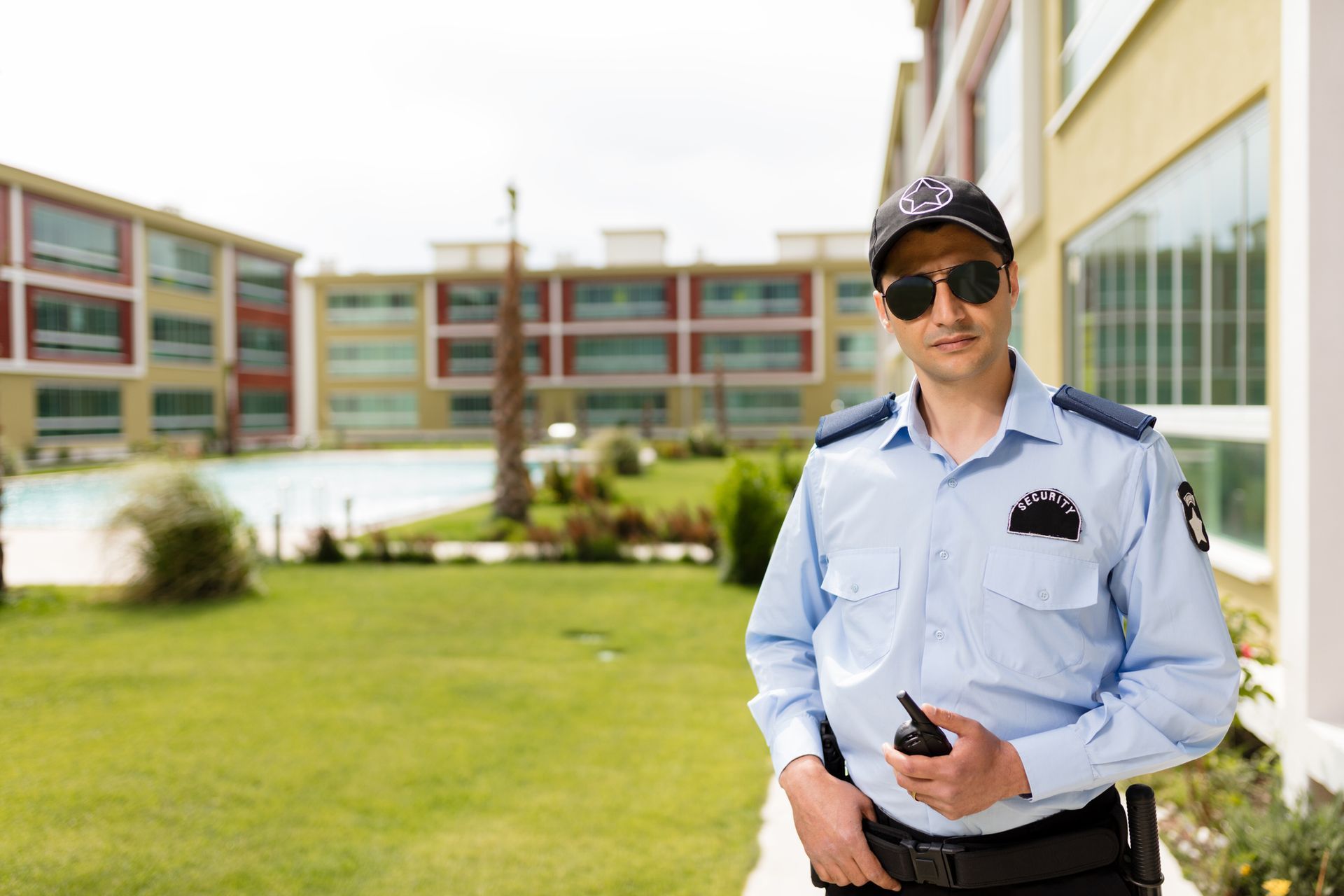 A security guard is standing in front of a building with a walkie talkie in his hand.