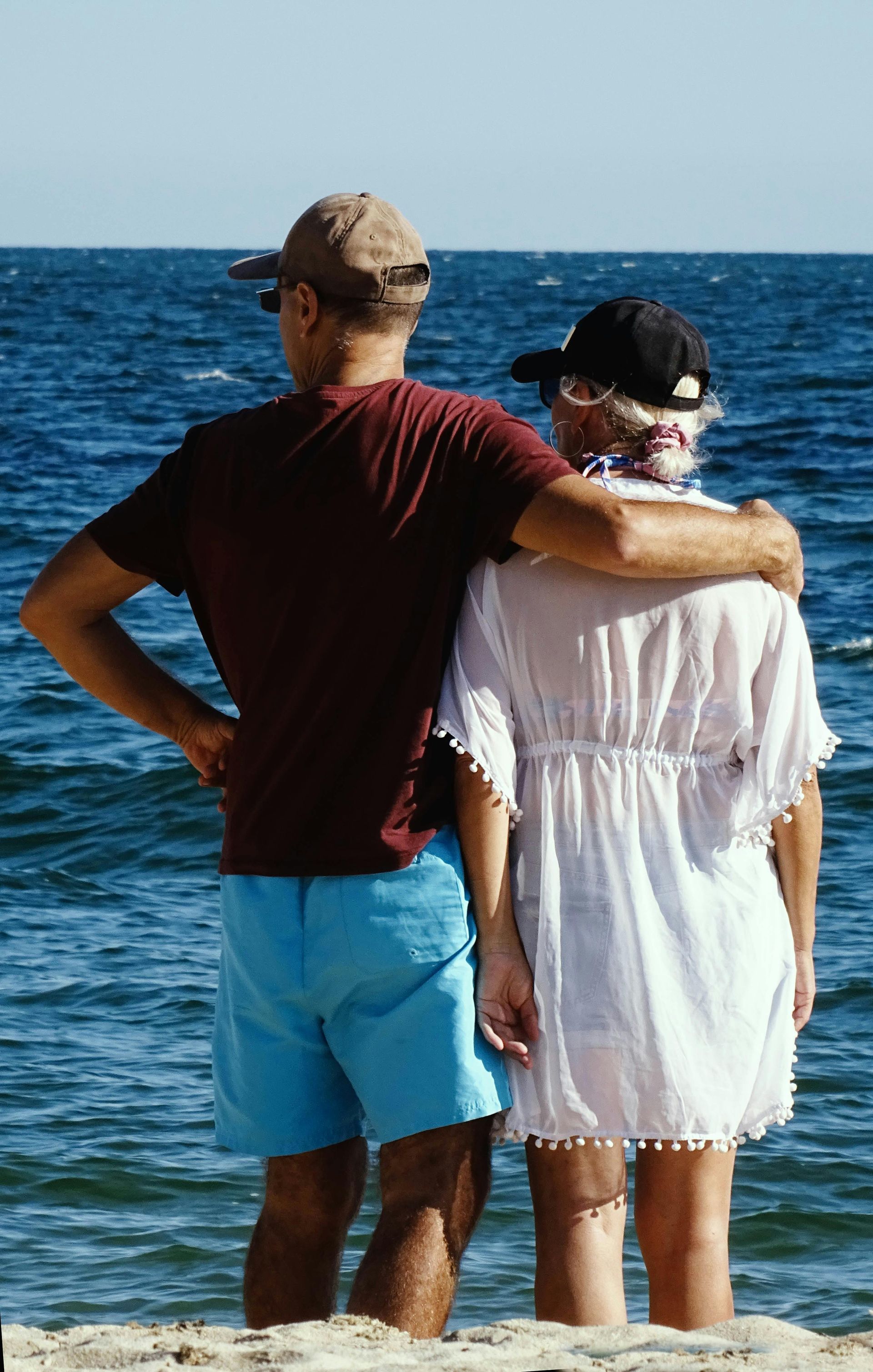 Two people standing by the sea, one with an arm around the other, facing the ocean