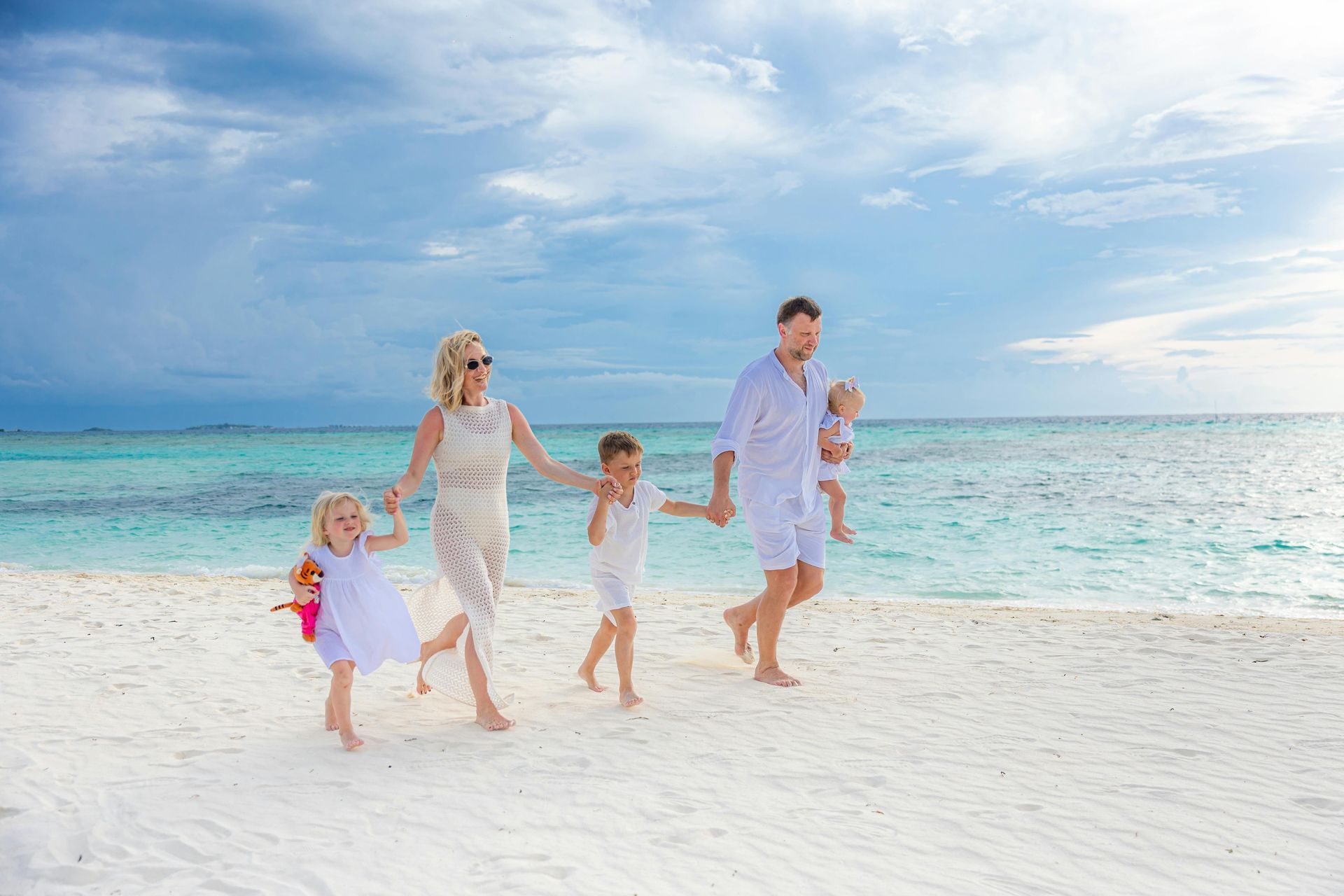 Family holding hands and walking on a bright white beach beside turquoise water under a blue sky