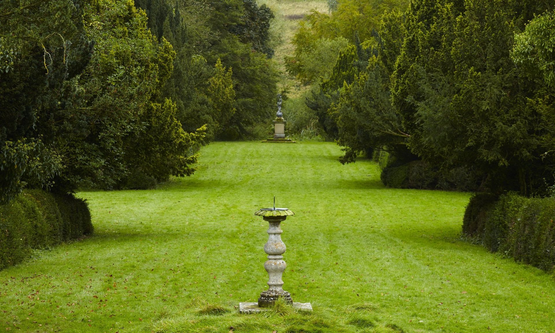 Lush green lawn pathway lined by trees, leading to a distant statue, with a sundial in the foreground.