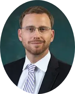 Professional headshot of a person wearing glasses, a dark blazer, and a striped tie against a dark green background.