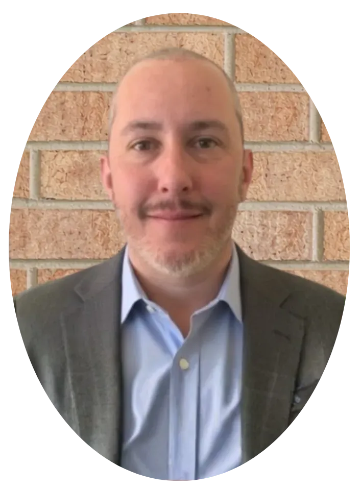 A person with short hair and a light beard, wearing a blue shirt and grey blazer against a brick wall background.