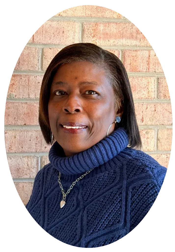 A person smiling, wearing a blue turtleneck sweater and a silver necklace, set against a light brick wall background.