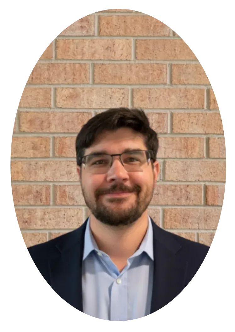 A person with dark hair, a beard, and glasses wearing a blue dress shirt and blazer against a brick wall background.