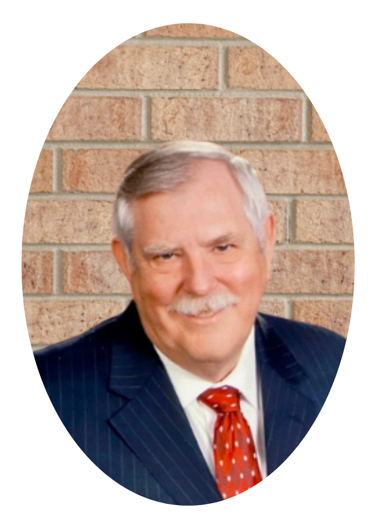 A smiling man with a mustache, wearing a navy blue pinstripe suit and red patterned tie, posing against a brick wall.
