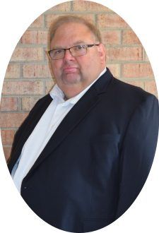 A person wearing glasses, a white collared shirt, and a dark blazer, positioned against a brick wall background.