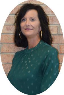 A person with dark hair and gold hoop earrings wears a green polka-dot top, posing against a brick wall.