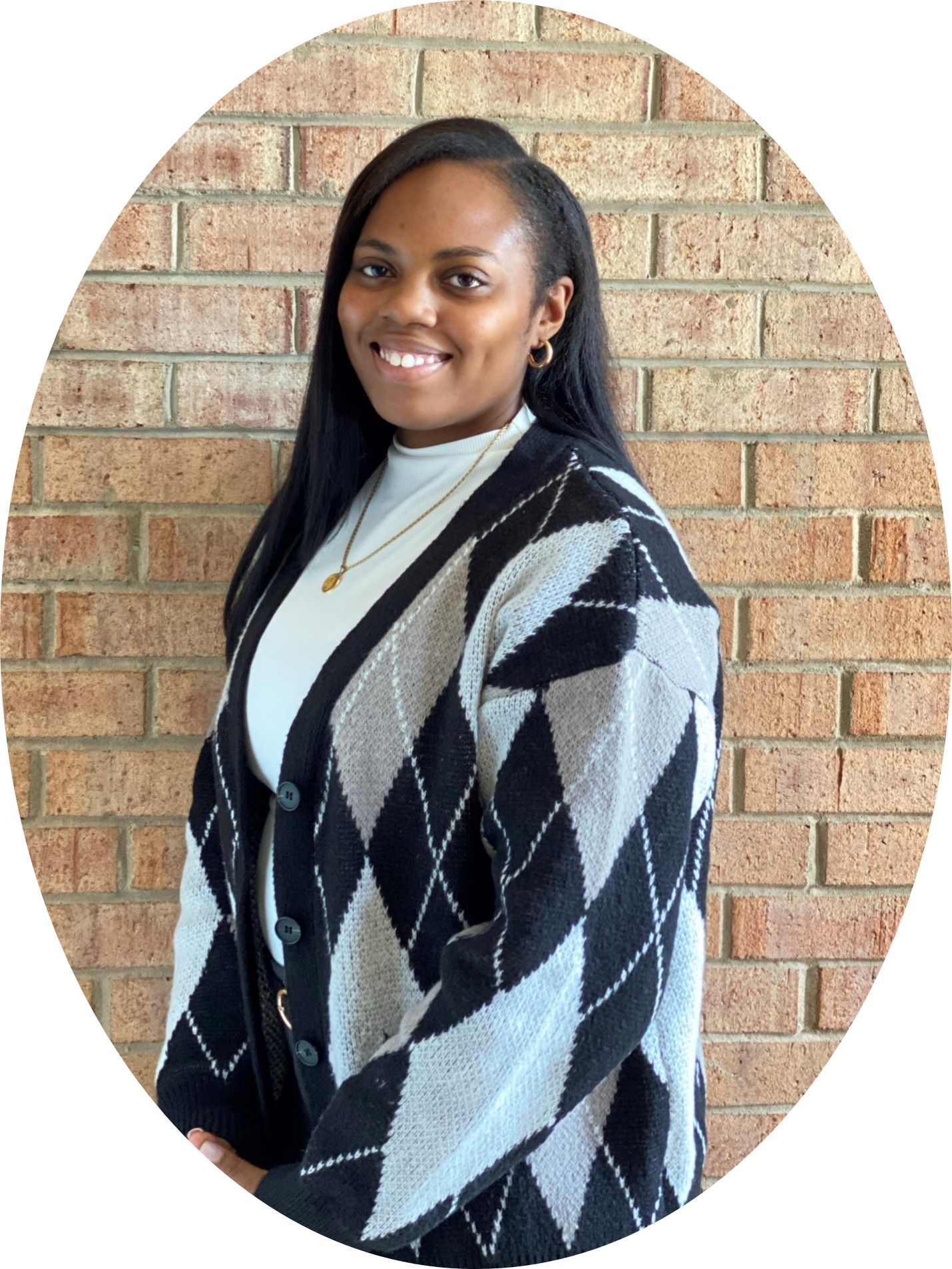 A person with long dark hair smiling, wearing a black and gray argyle cardigan over a white shirt, against a brick wall.