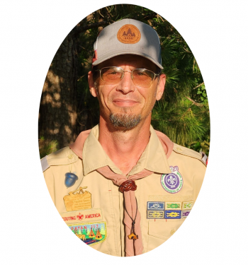 A person wearing a tan Boy Scouts of America uniform and cap, smiling against a background of trees.