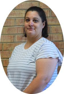 A person with dark hair wears a white shirt with horizontal black stripes, standing in front of a tan brick wall.