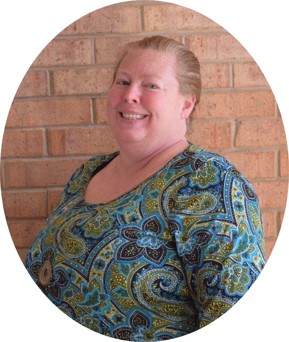 A person smiling in front of a brick wall, wearing a blue and green paisley patterned top.