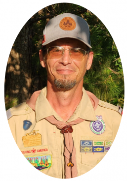 A person wearing a tan Boy Scouts of America uniform and cap, smiling against a background of trees.