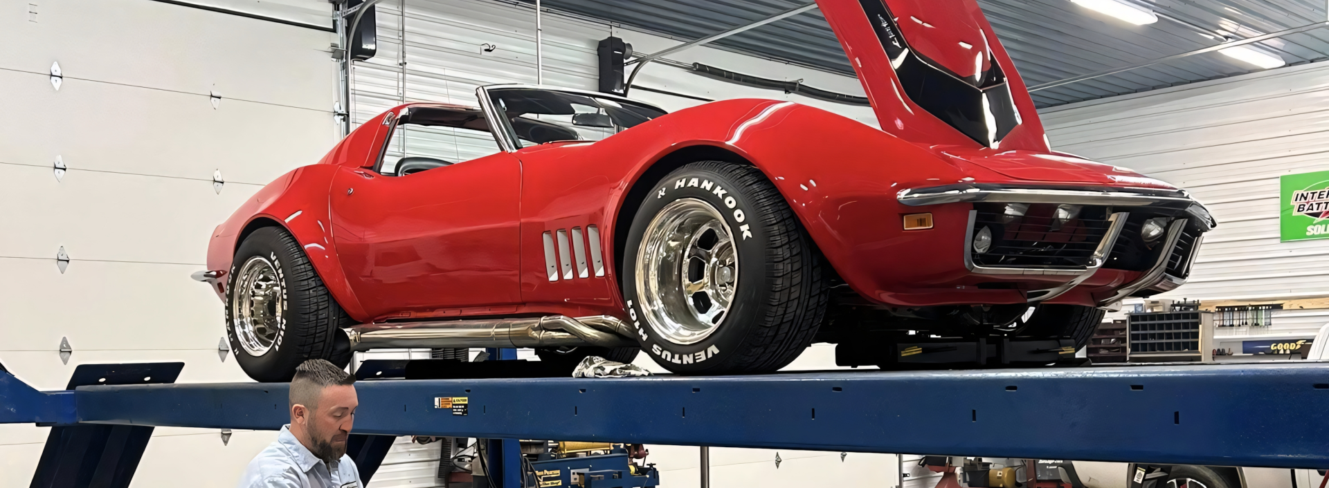 Red vintage Corvette on a car lift in a garage with the hood open, a man looking at the car | Chip Ragans Auto, LLC