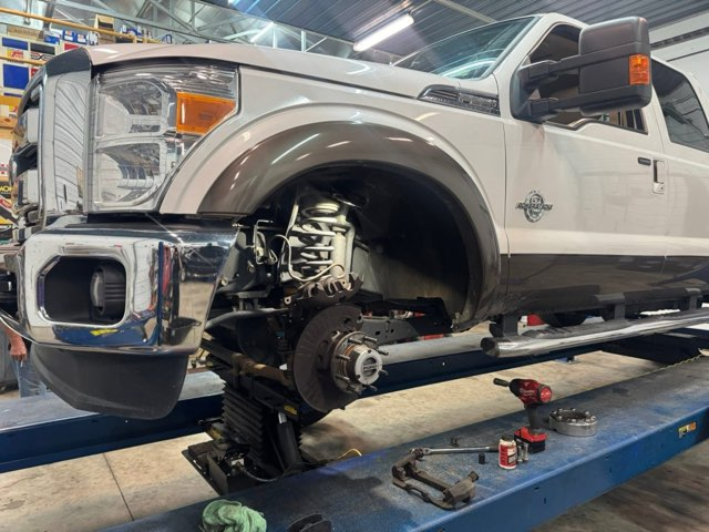 White truck on a lift with front wheel removed, mechanic working in a garage | Chip Ragans Auto, LLC
