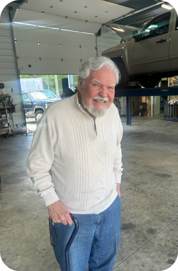 Man with white hair and beard smiles in a garage. He wears a cream sweater and jeans | Chip Ragans Auto, LLC