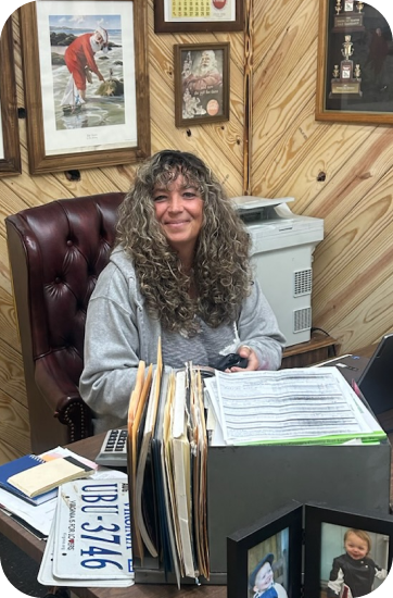 Woman seated at a desk, smiling, with files, license plate, and photos in an office setting | Chip Ragans Auto, LLC