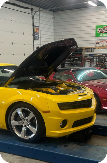 Yellow Camaro with open hood on a lift inside a repair shop. A red car is partially visible in the background | Chip Ragans Auto, LLC
