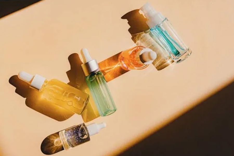 Skincare products in colorful glass bottles with droppers, angled on a beige surface with sunlit shadows.