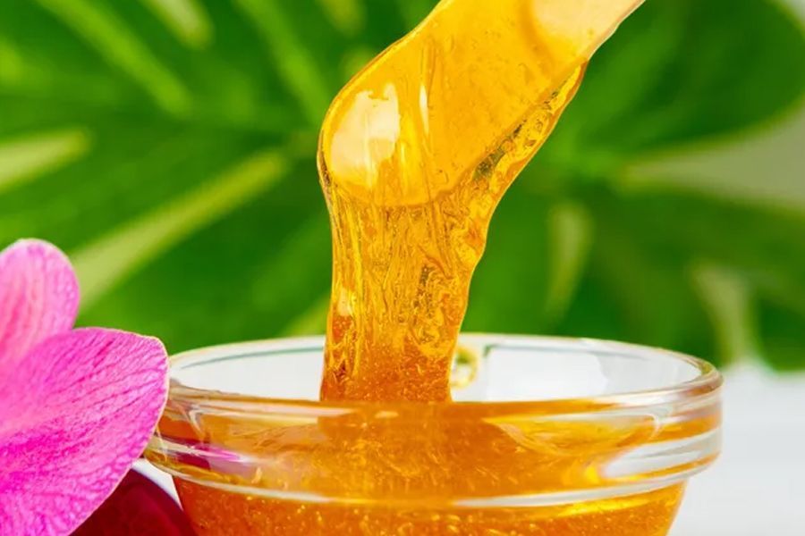 Wax on wooden applicator dripping into a small glass bowl; pink flower and green foliage in background.