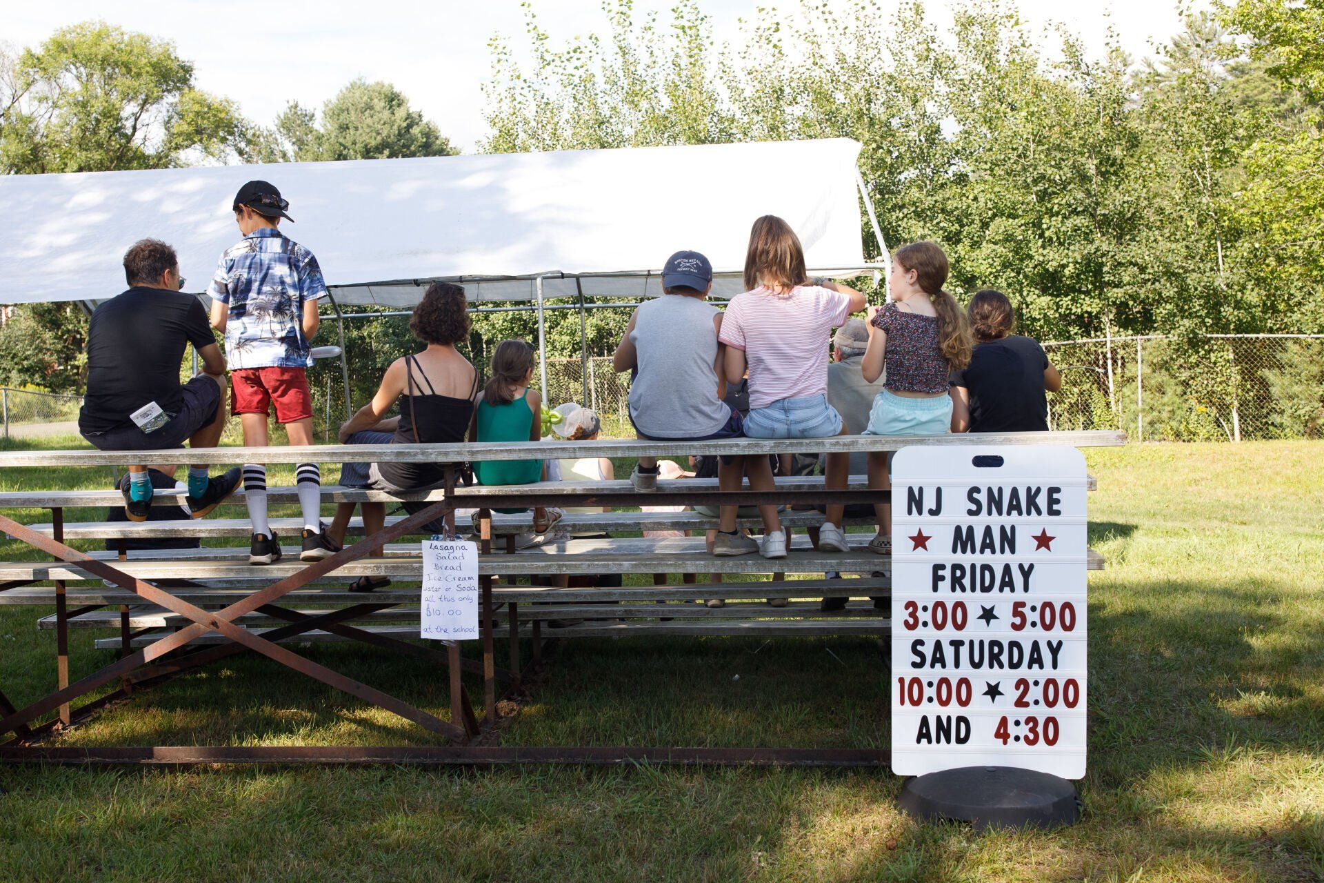 A group of people sitting on a bleacher with a sign that says nj shake man friday saturday and 4:30