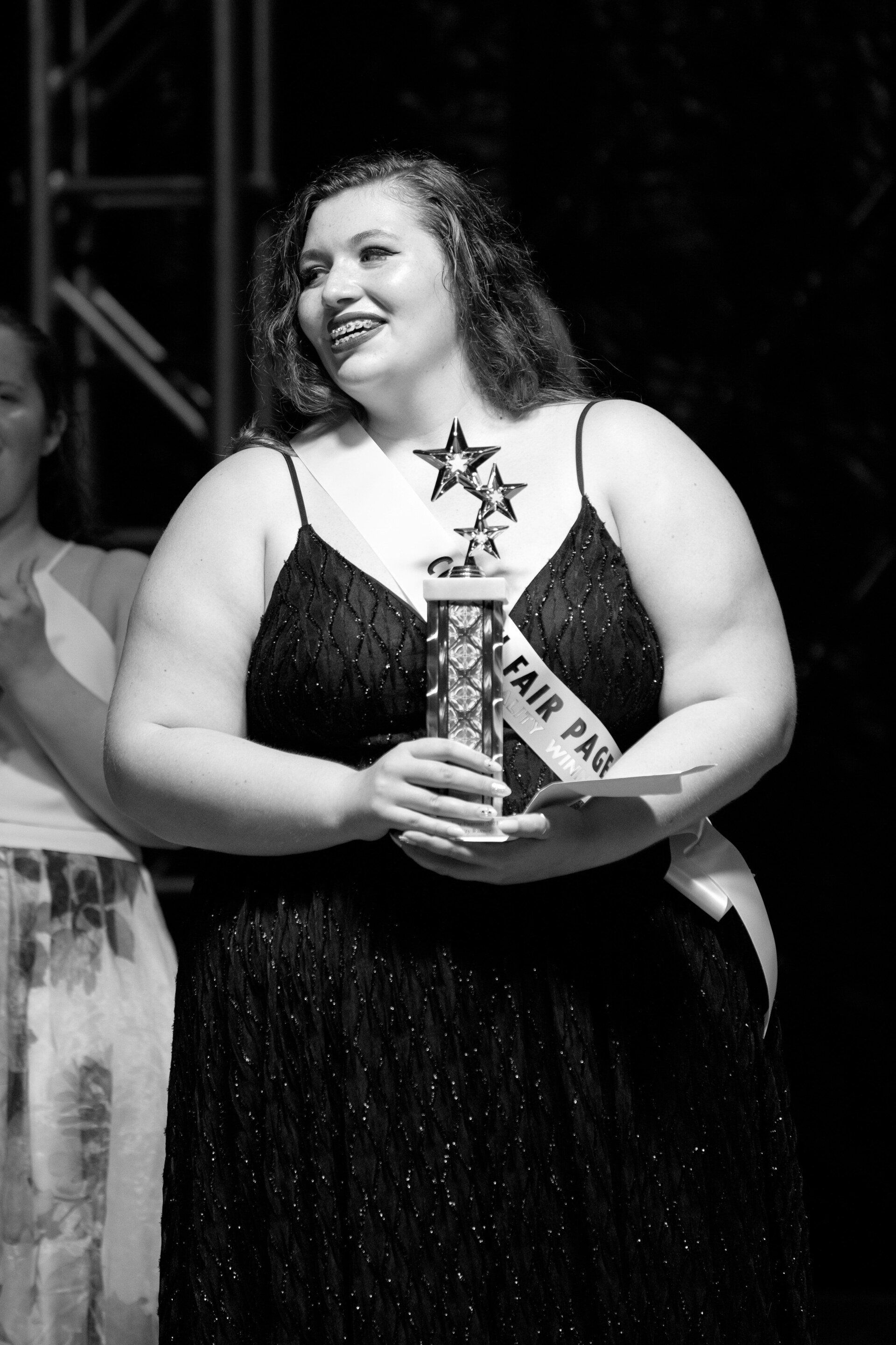 A woman in a black dress is holding a trophy that says fair pageant