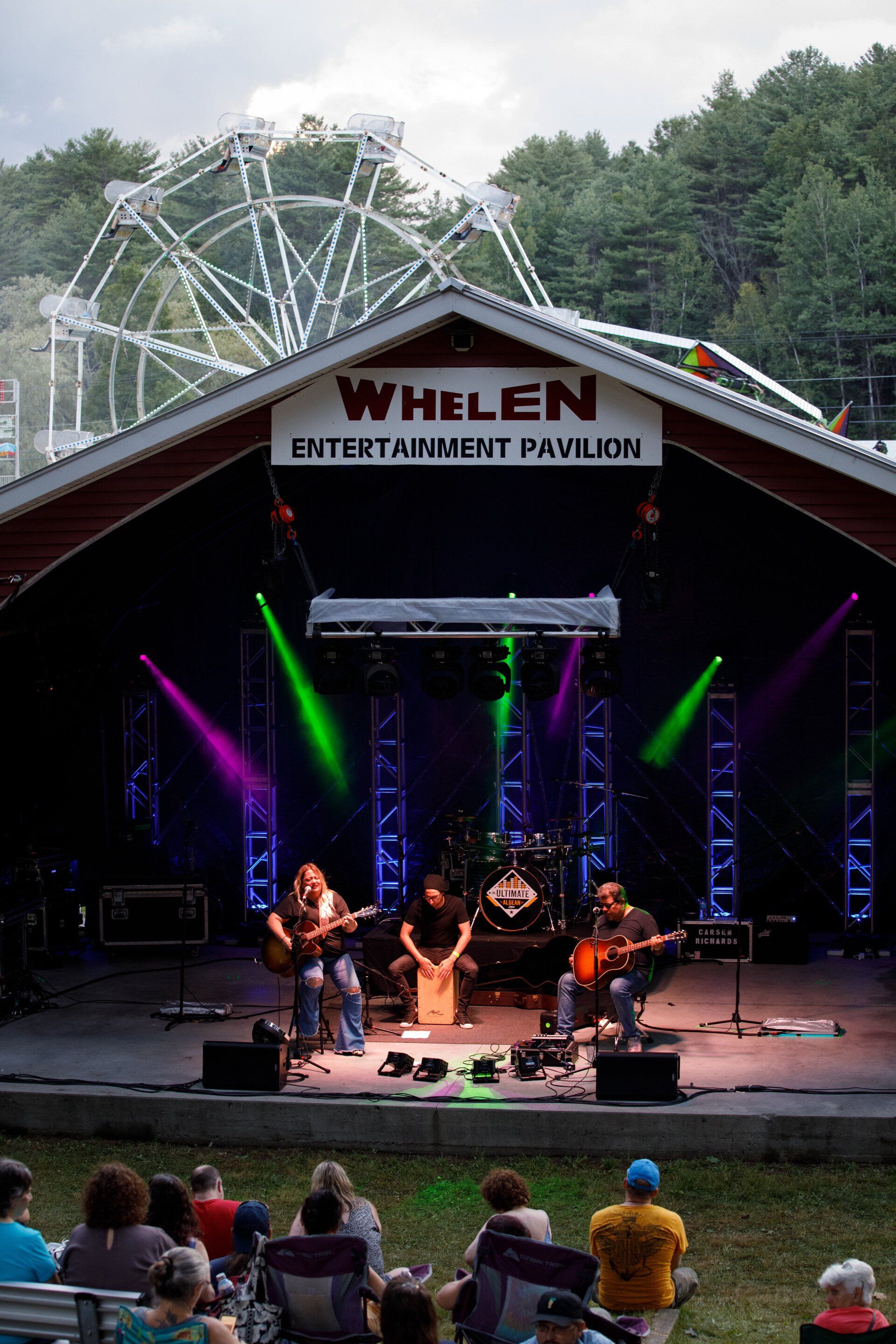 A group of people are sitting in front of a stage with a ferris wheel in the background.