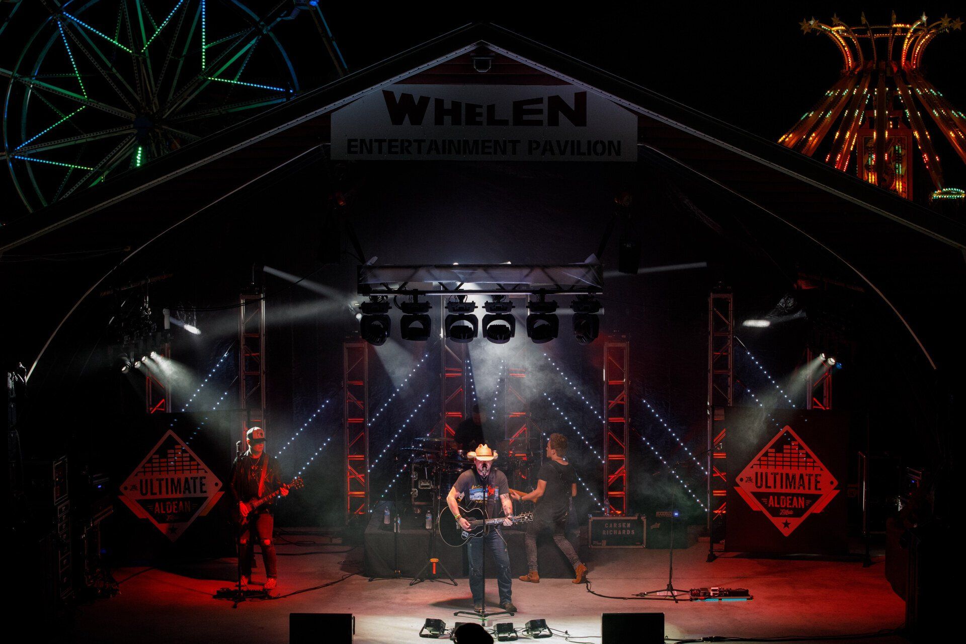 A man is playing a guitar on a stage in front of a ferris wheel.