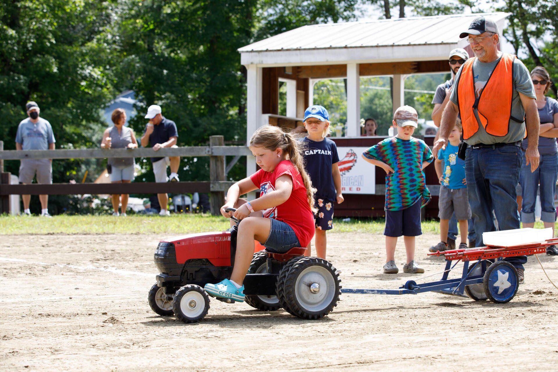 A little girl is riding a toy tractor pulling a wagon.