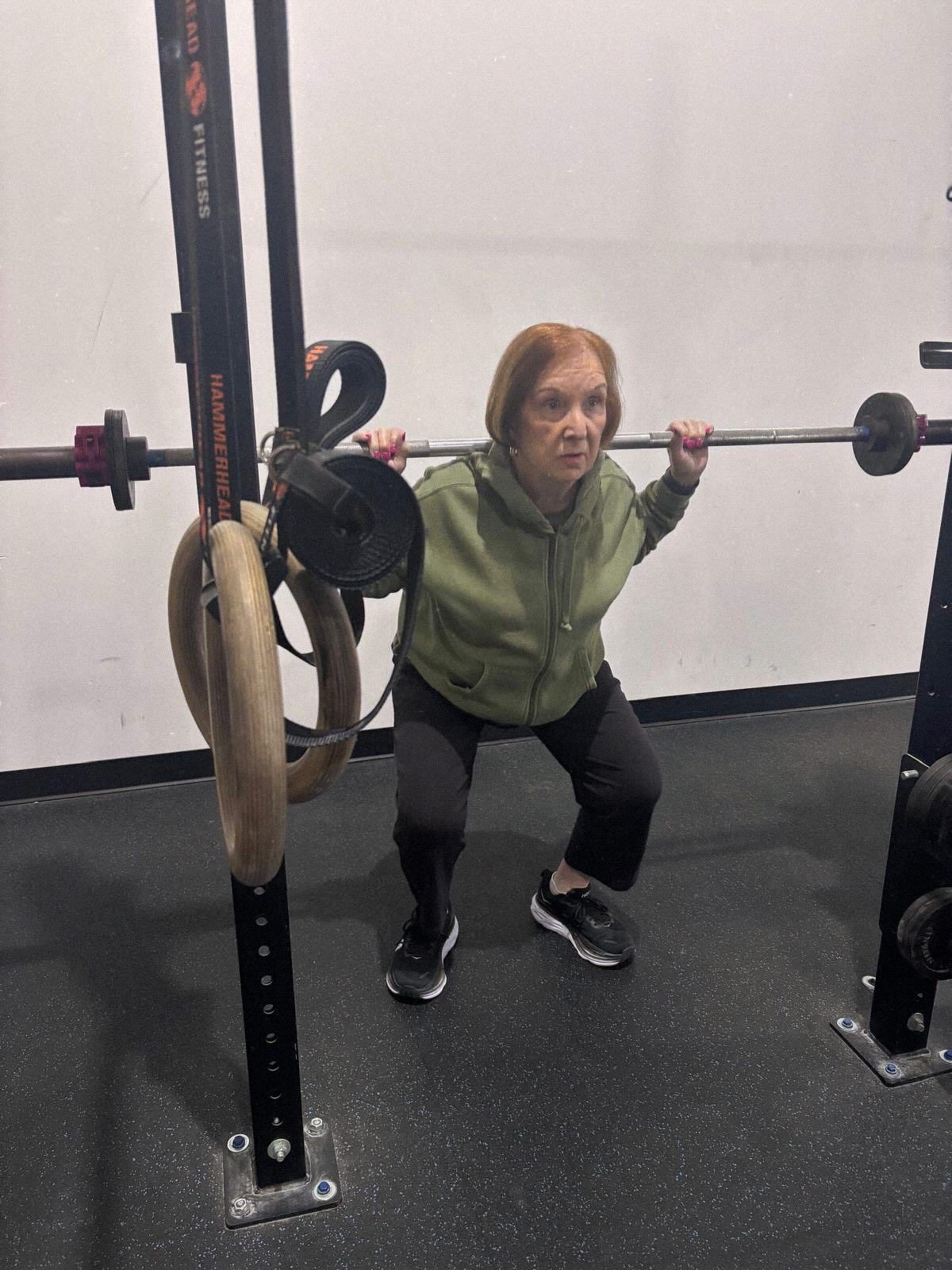 An older person performs a barbell squat in a gym. They wear a green hoodie and black pants.