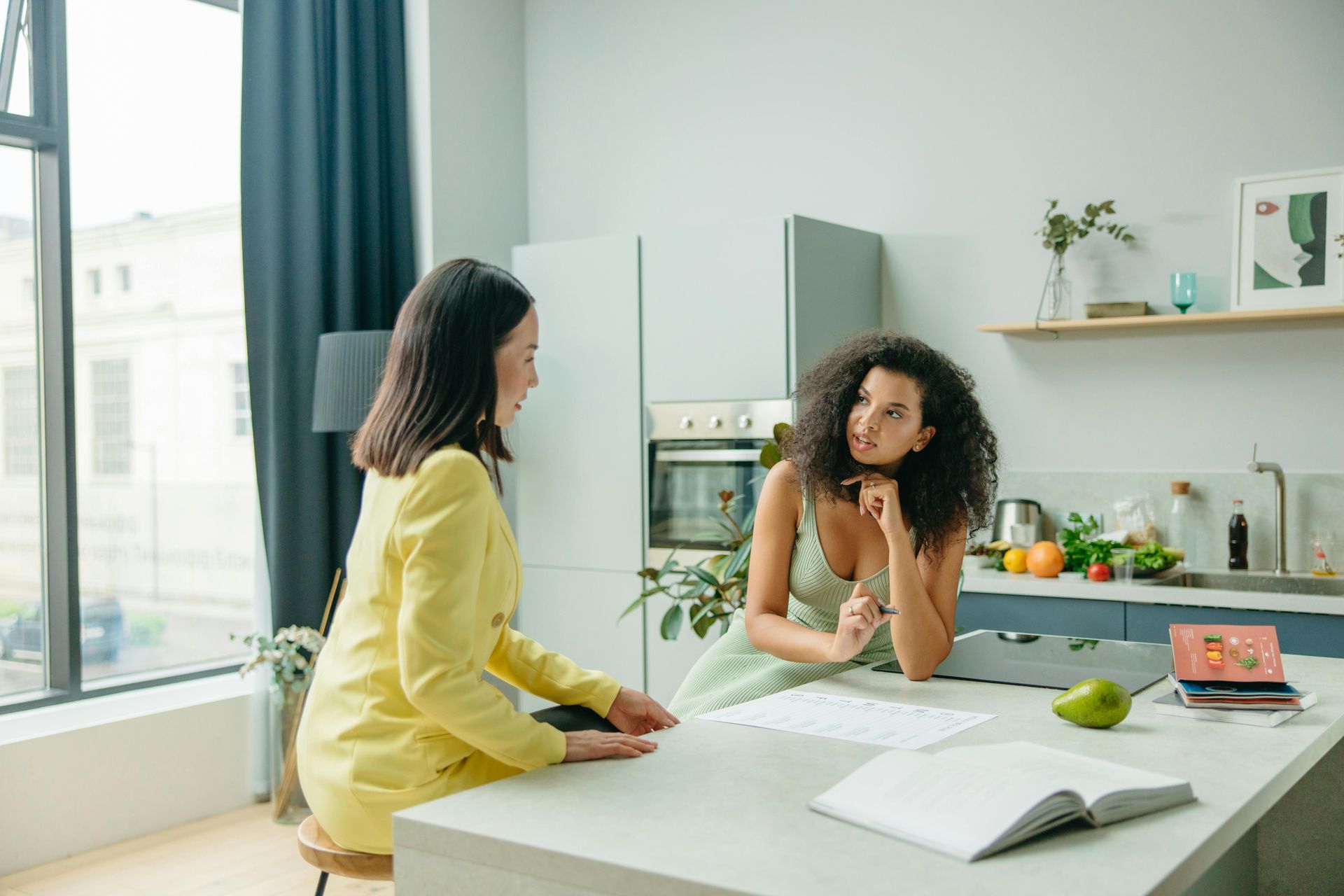 Two women in a bright kitchen: one in yellow jacket, the other in a floral dress, reviewing papers at a counter.