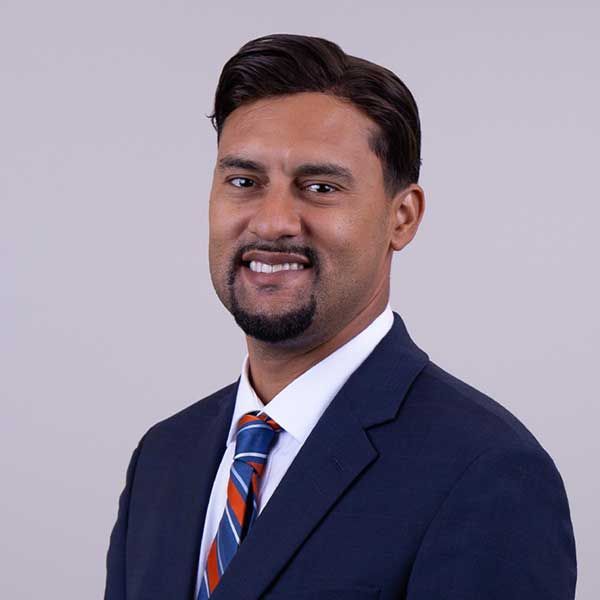 Man in navy suit, smiling, with a blue and orange tie, against a light gray background.