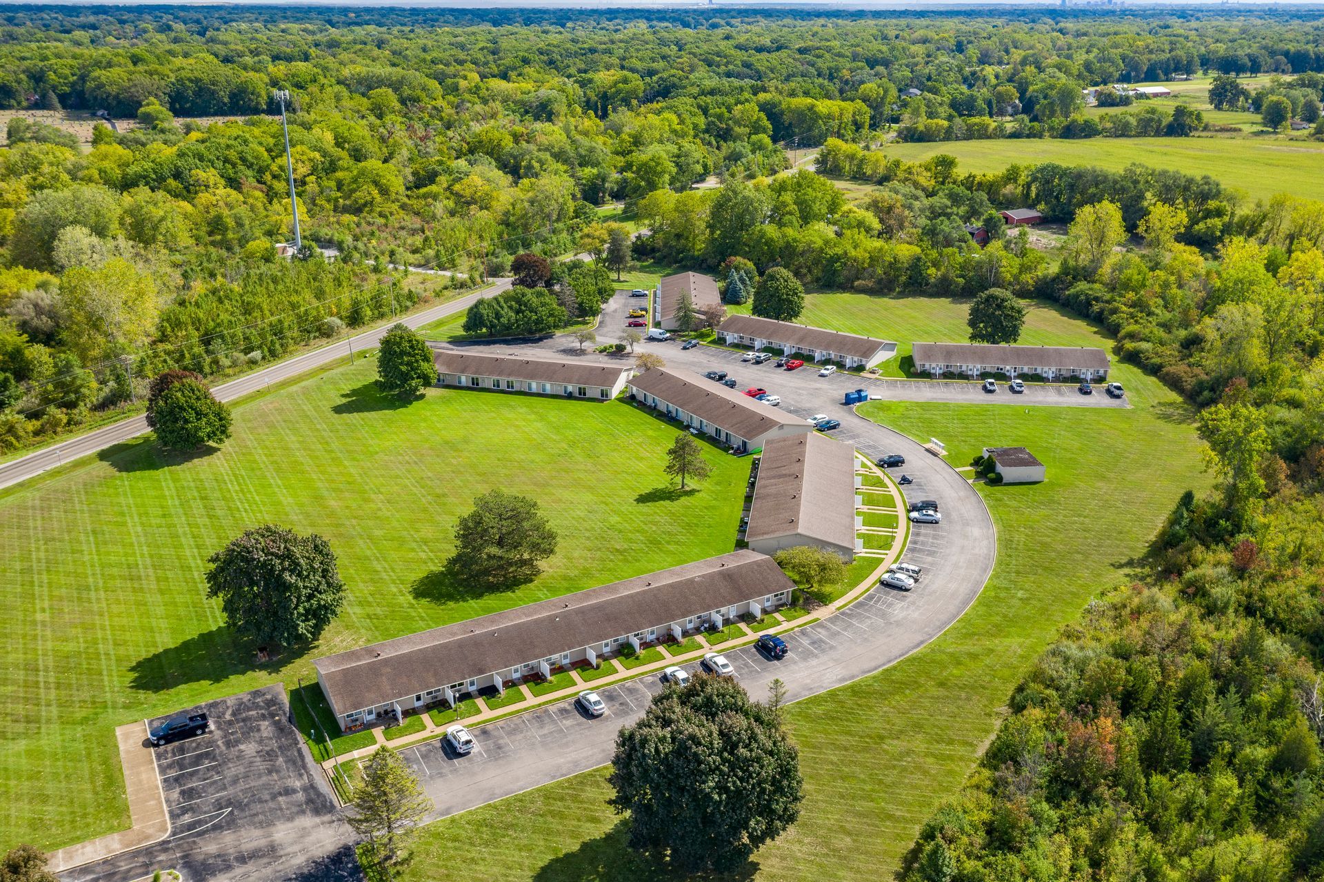 Aerial view of apartment complex with brown roofs and a circular driveway on a grassy landscape surrounded by trees.