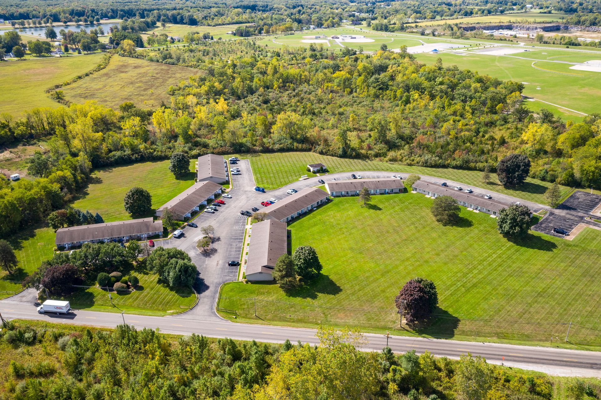 Aerial view of a complex of tan, single-story townhomes with green lawns and trees, surrounded by fields and a wooded area.