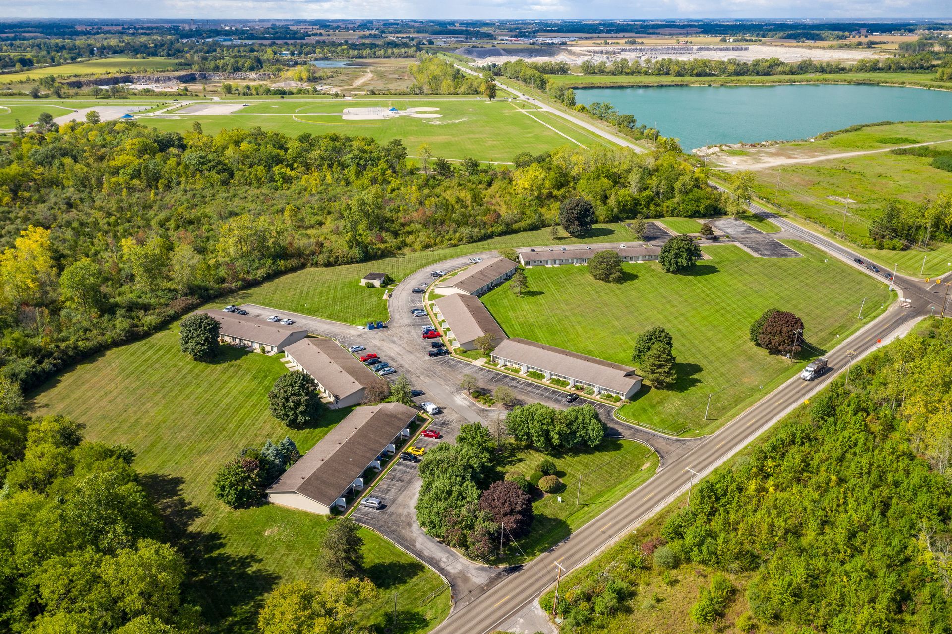 Aerial view of tan buildings in a grassy area, surrounded by trees, roads, and a lake.