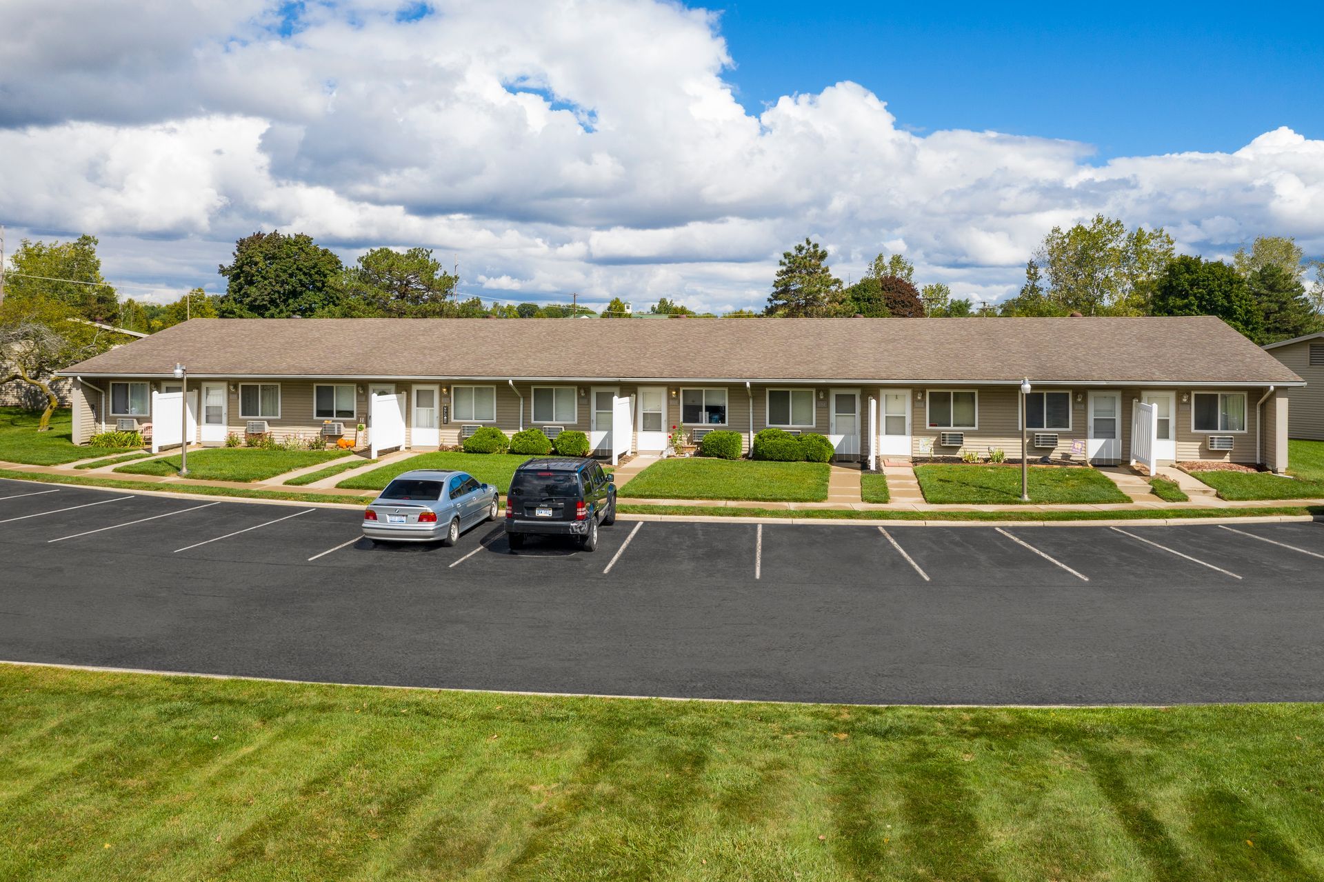 Apartment building with cars parked in front. Gray siding, brown roof, and green lawn. Cloudy sky.