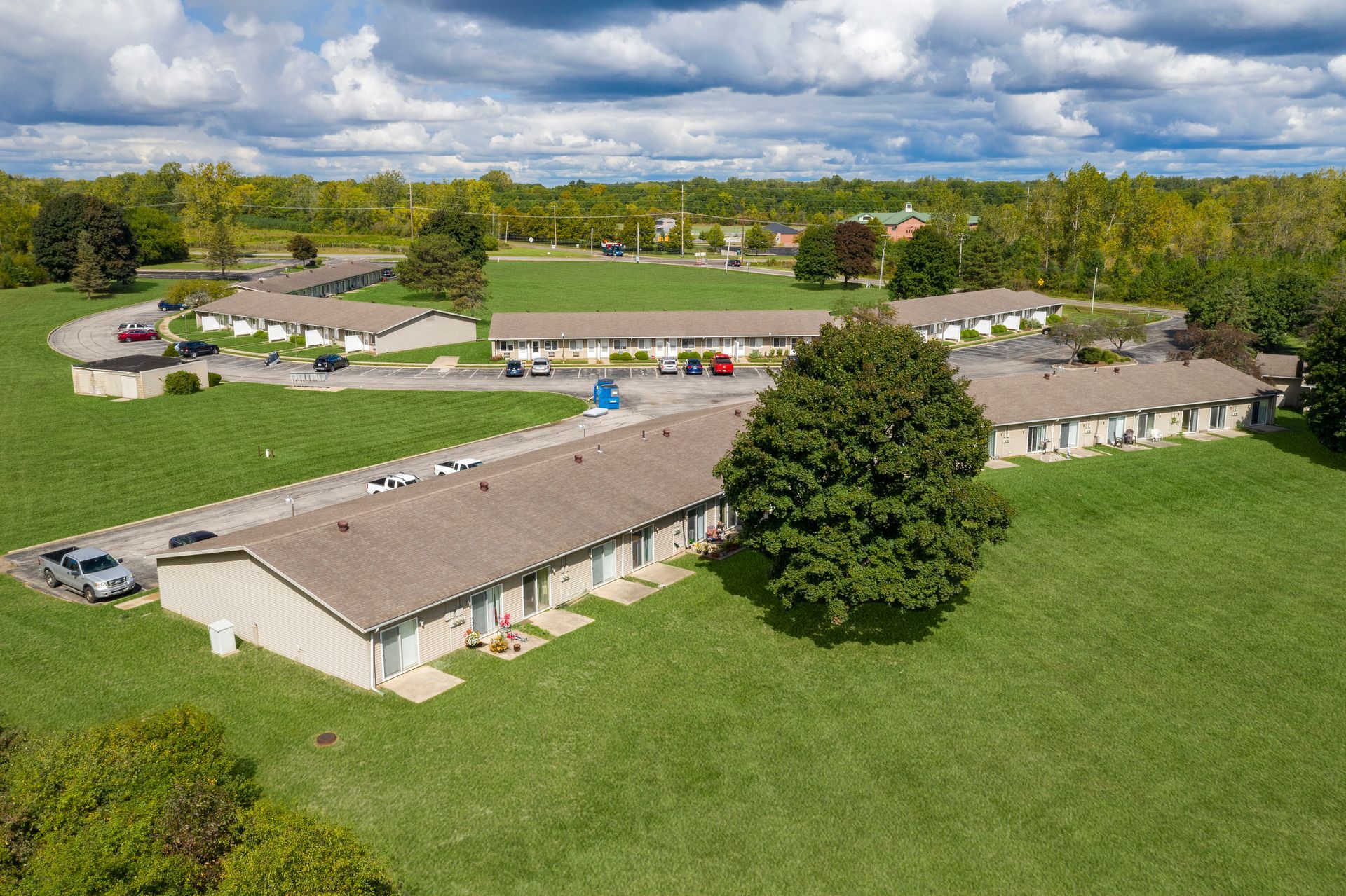 Aerial view of beige apartment buildings with brown roofs on a green lawn, surrounded by trees.
