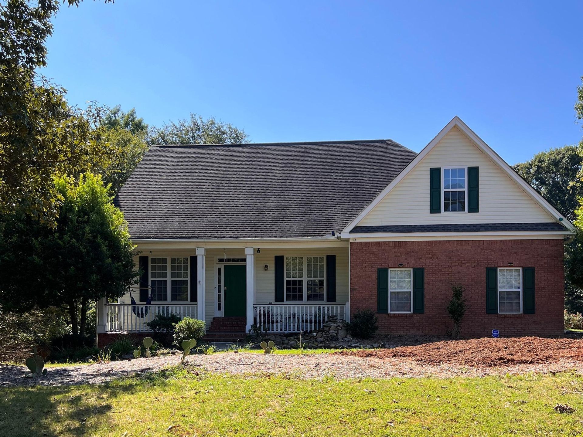 A one-story brick house with a white front porch, green shutters, a gable roof, and a lawn under a clear blue sky.
