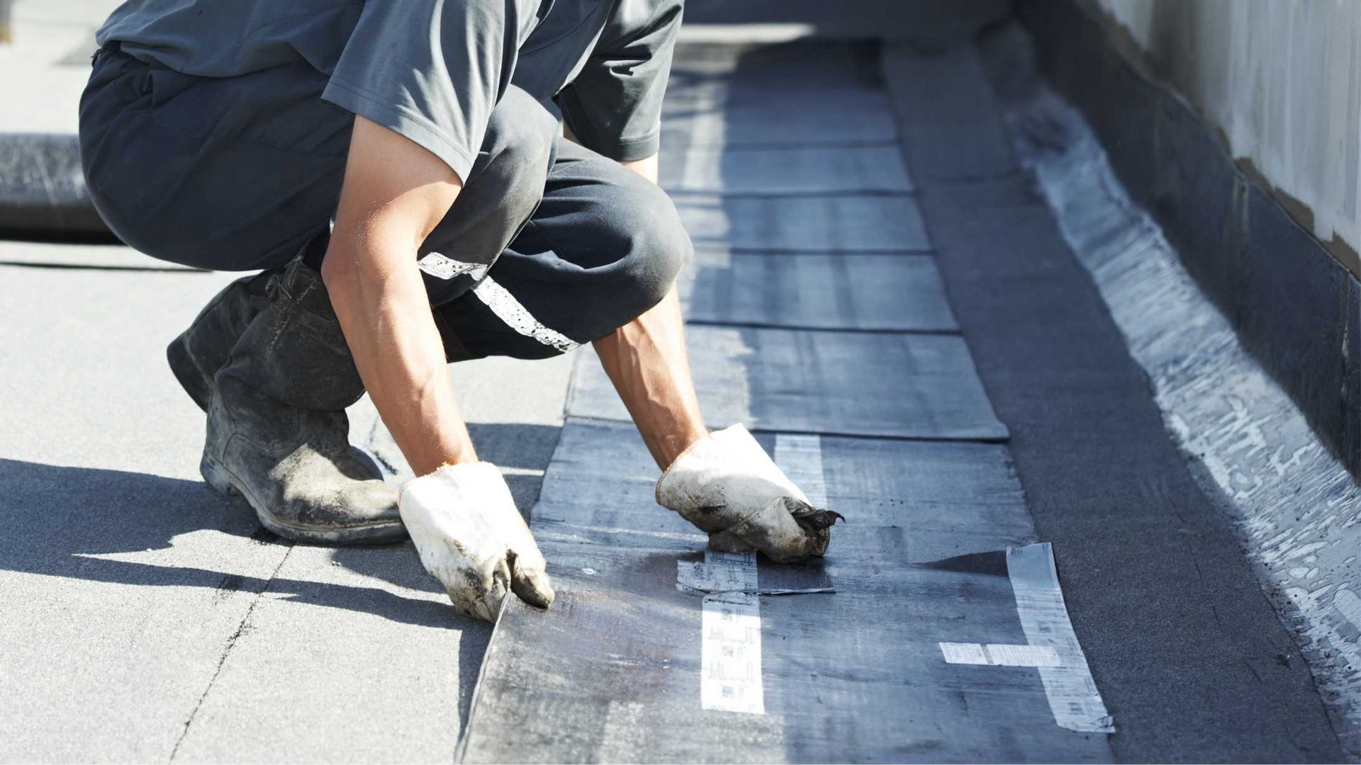 A person wearing work gloves kneels on a flat roof, applying a strip of adhesive material to the surface.