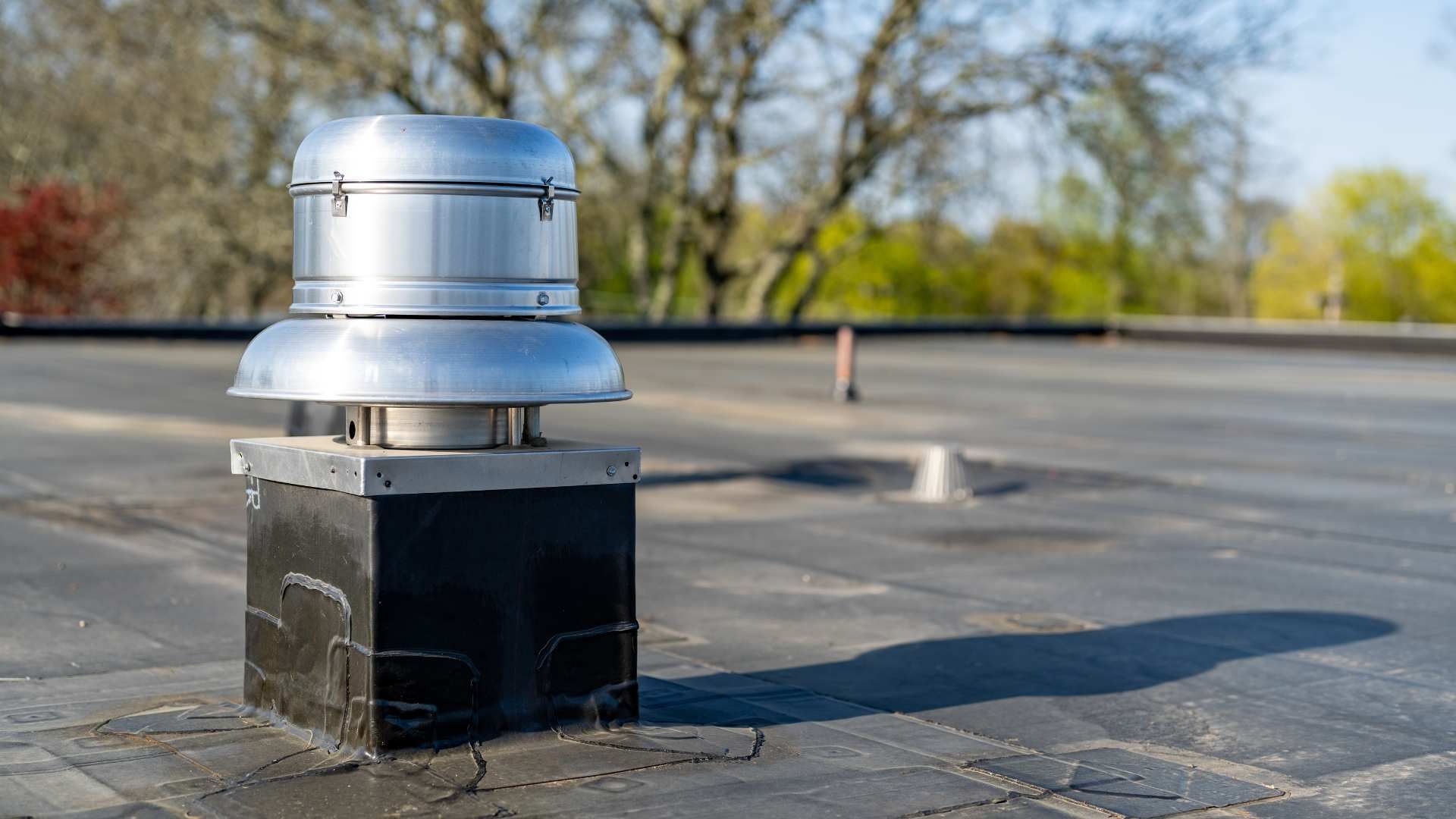 A silver metal roof vent mounted on a square black curb atop a flat roof with trees in the blurred background.