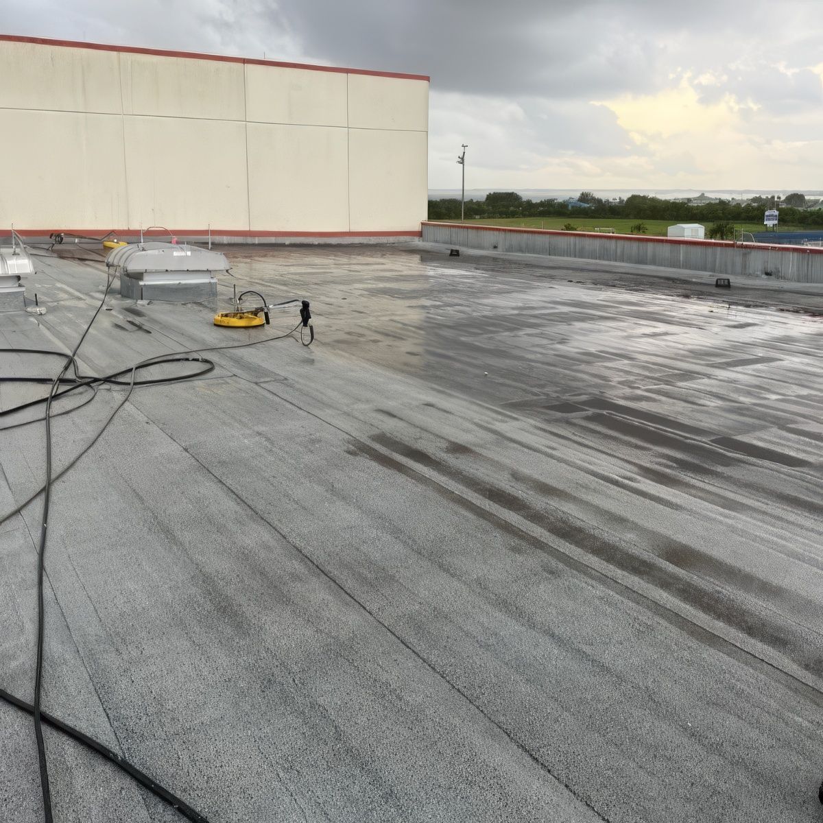 A wide, grey, flat commercial roof with moisture patterns, a yellow tool, and a beige wall under a cloudy sky.