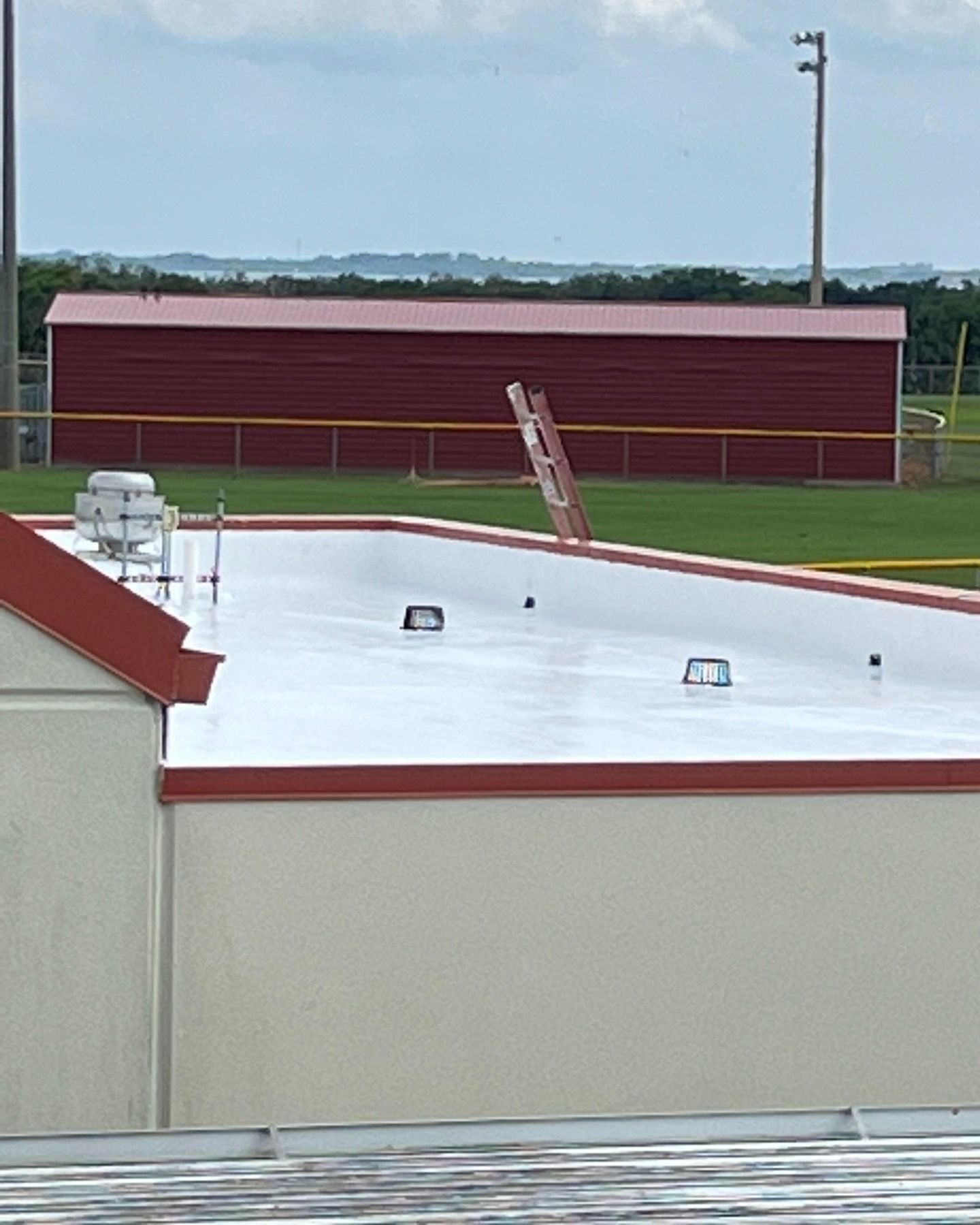 A white, flat roof with vents and a ladder leaning against the far edge, overlooking a field with a red building.