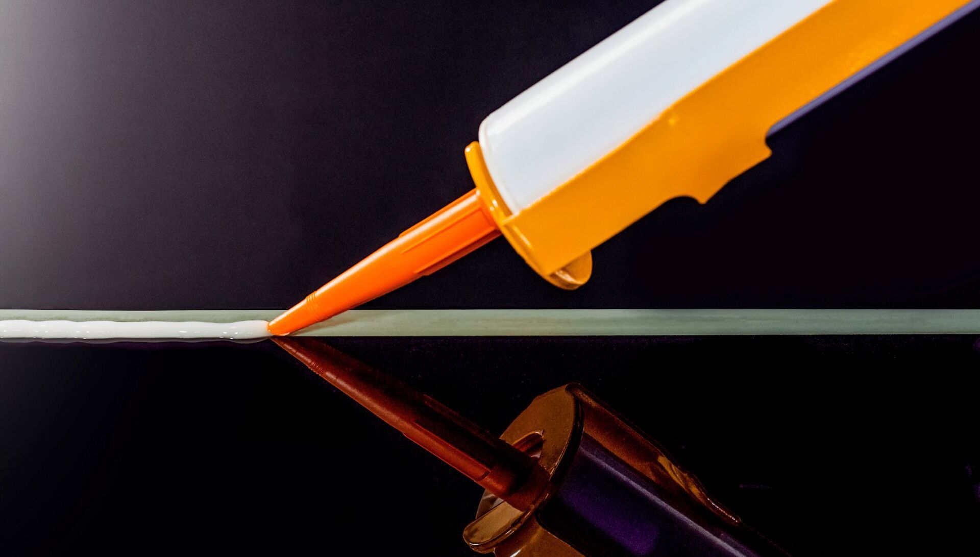Caulk gun dispensing a bead of caulk onto a reflective black surface.