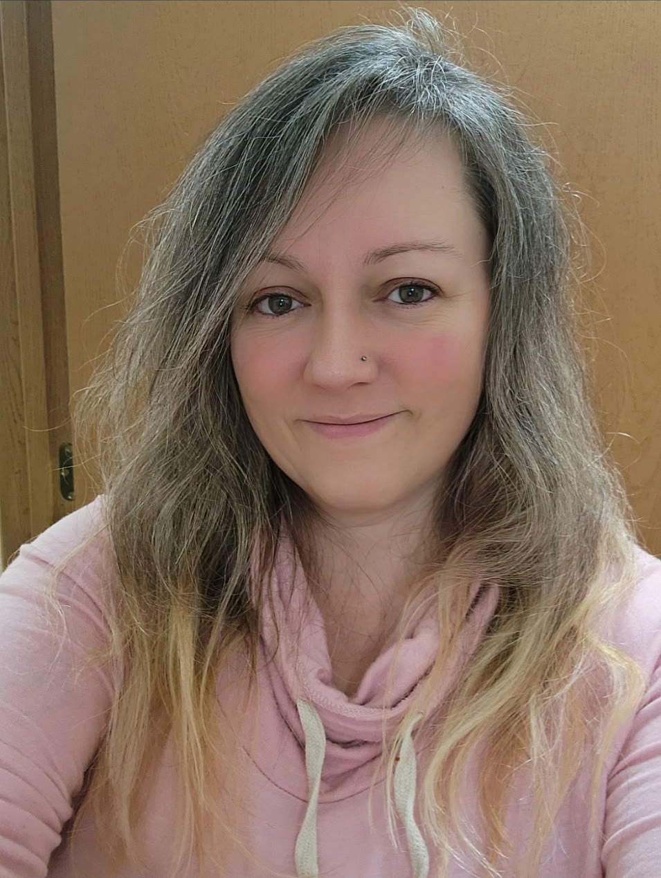 A person with shoulder-length, two-toned hair wearing a light pink, hooded cowl-neck top against a plain brown background.