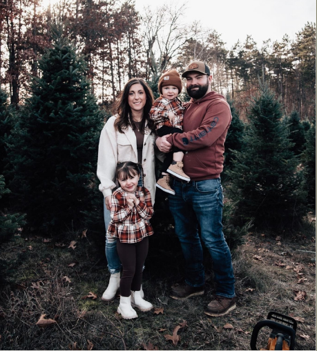 A family of four poses together at a Christmas tree farm, surrounded by evergreen trees under a soft, overcast sky.