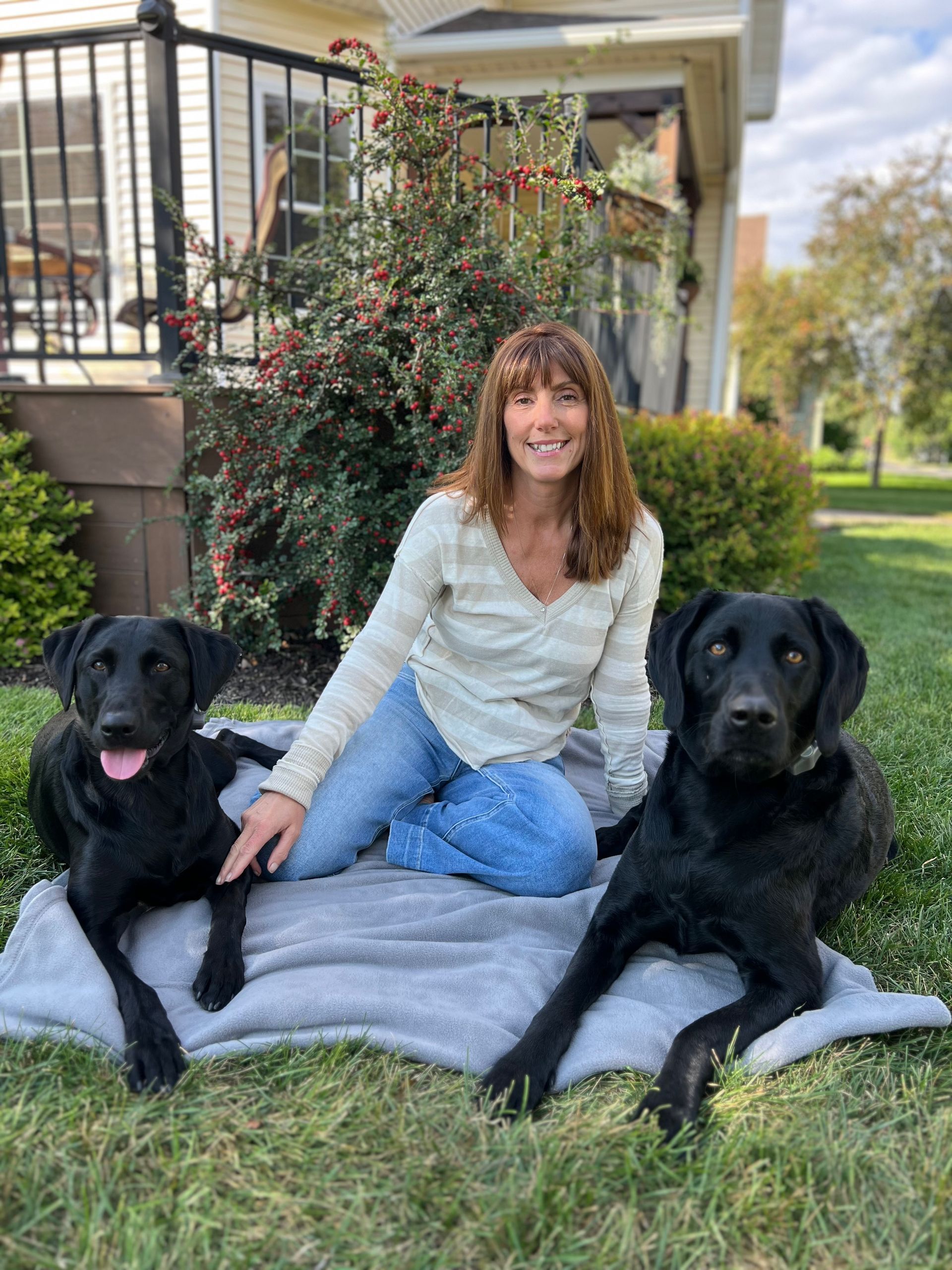 A person sitting on a blanket in a yard with two black dogs, one with its tongue out, next to a house and a large shrub.