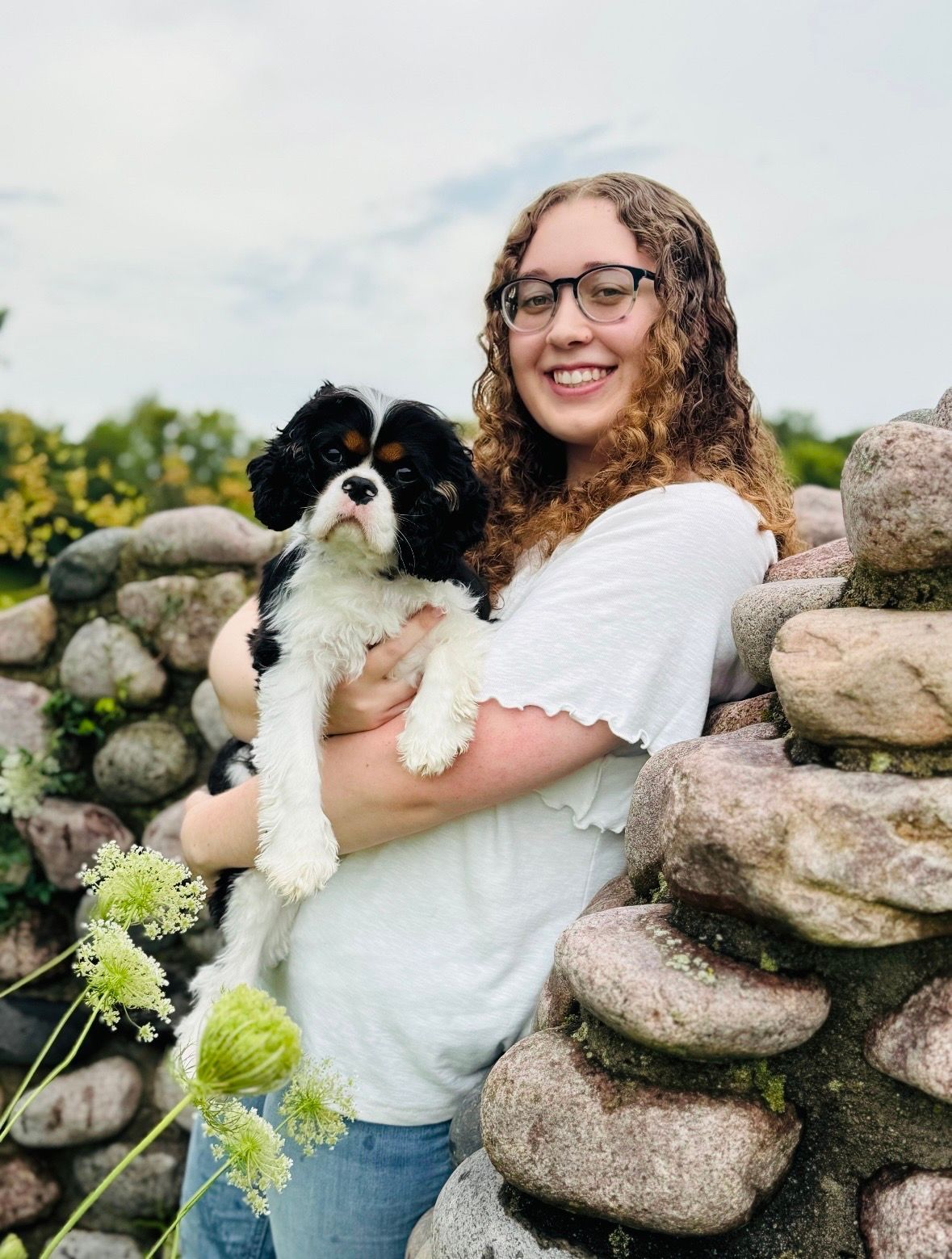 A person with curly hair and glasses holds a small, black-and-white Cavalier King Charles Spaniel by a stone wall.