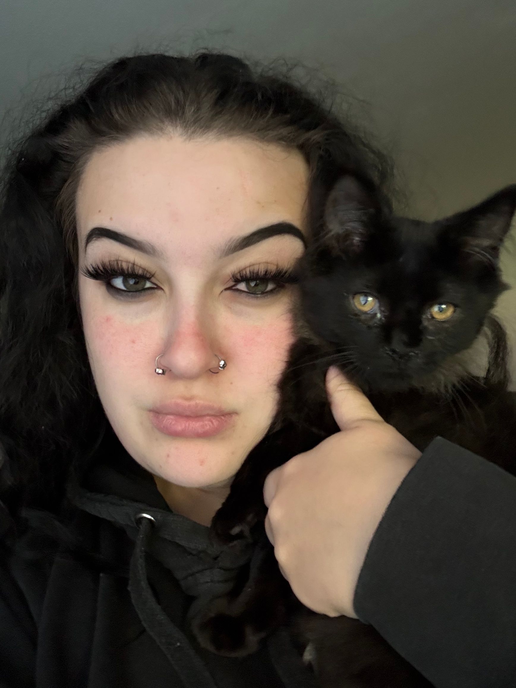 A person with dark hair and a septum piercing holding a small, black kitten against their chest.