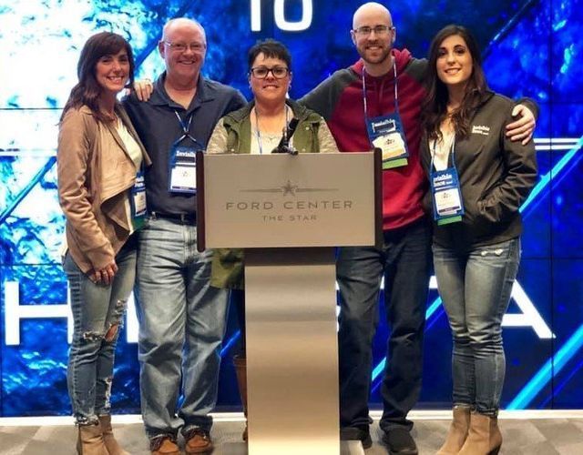Five people stand smiling behind a Ford Center podium in front of a large, blue, digital screen.