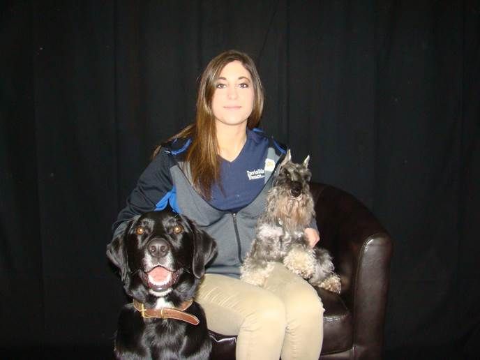 A person sits in a chair in front of a black curtain, flanked by a black dog on the left and a gray terrier on the right.